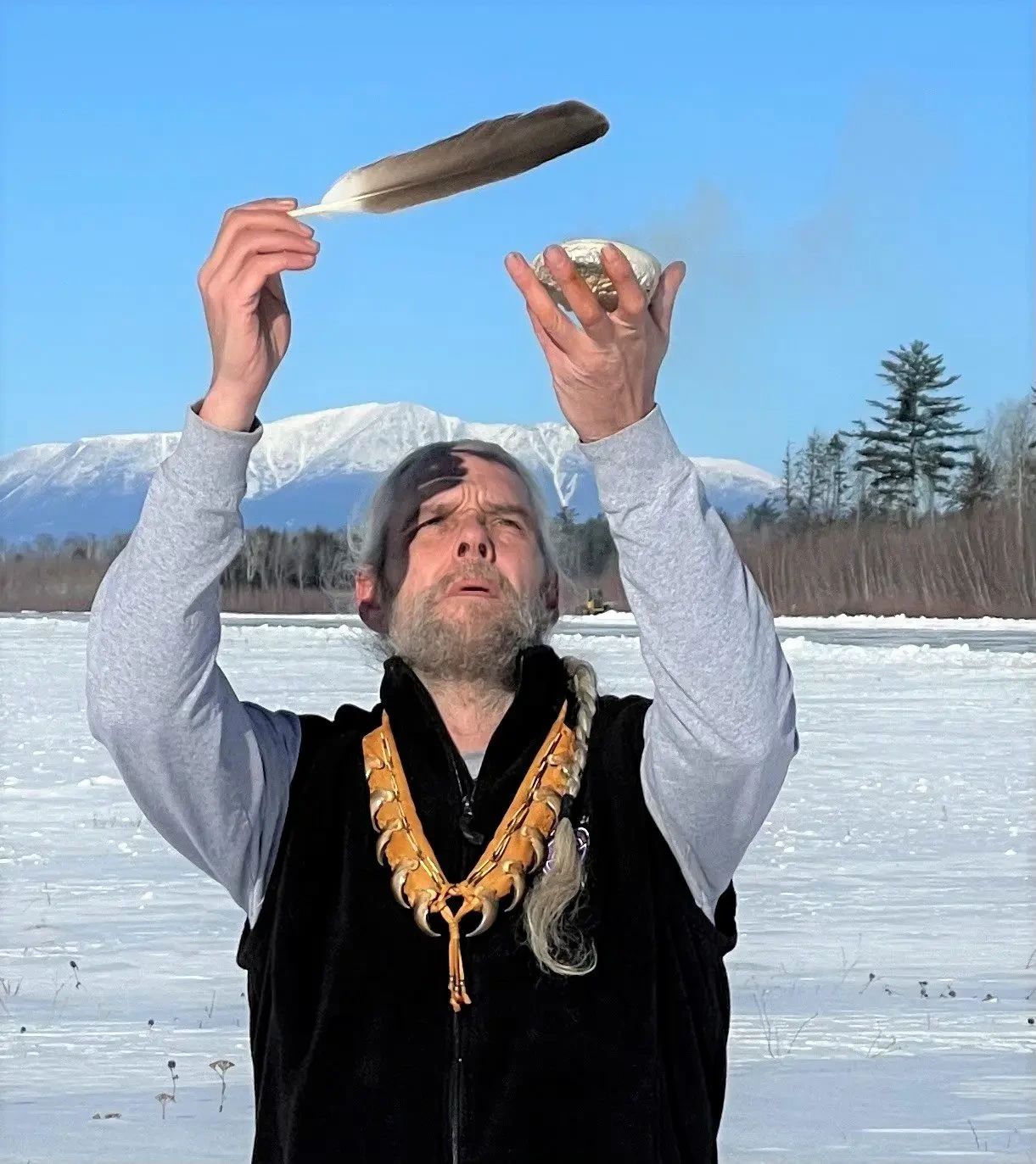 Man holds feather and wrapped item overhead while standing on frozen lake with mountains and evergreen trees behind him.
