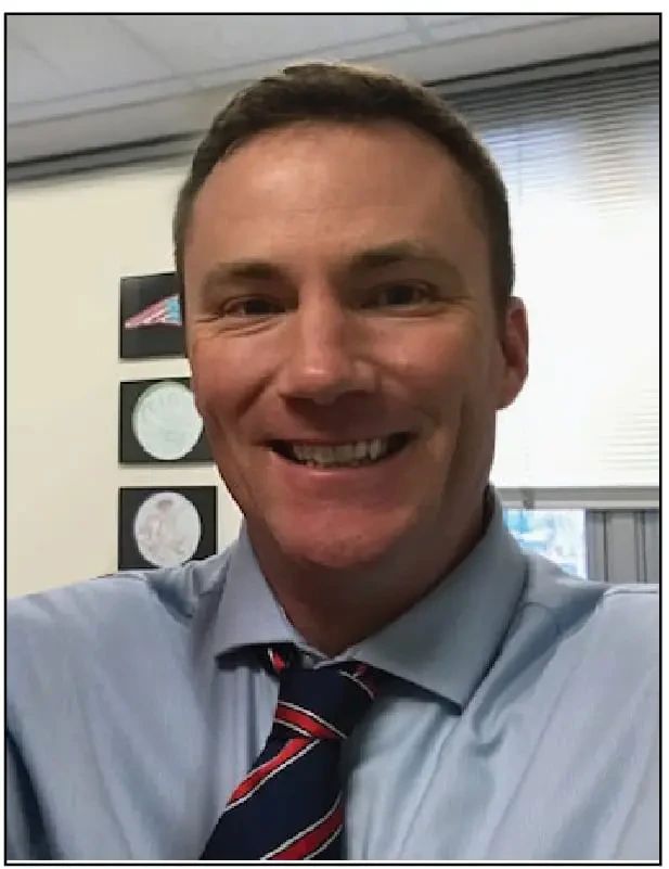 A smiling man wearing a light blue dress shirt and a navy blue necktie with red and white stripes.