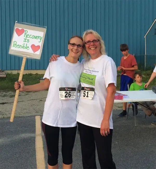 Two smiling women in white t-shirts with race bibs stand together; one holds a sign that says Recovery is Possible.