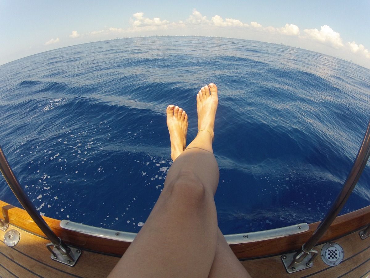 A person’s legs resting on the edge of a wooden boat deck, overlooking the open ocean.