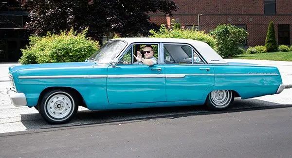 A man wearing sunglasses waves from the driver’s seat of a vintage teal sedan parked on an asphalt road.