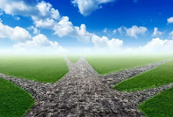 A stone path forks into three directions across a green field under a blue sky with clouds.