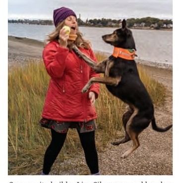 A woman in a red jacket holding a tennis ball as a dog jumps toward it on a beach.
