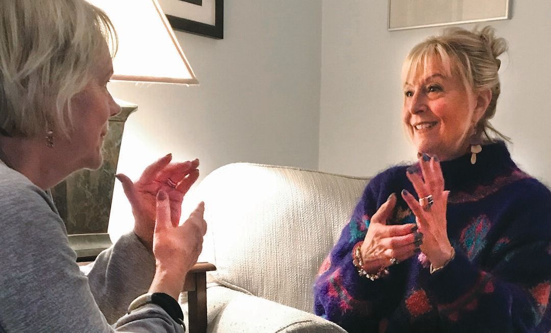 Two women sit facing each other on a couch, smiling and gesturing with their hands as they have a lively conversation about self-care in a cozy, warmly lit room.