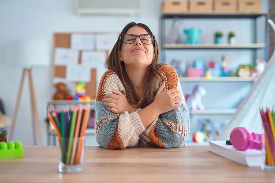 A smiling woman wearing glasses and a sweater sits at a desk, hugging herself with her eyes closed.
