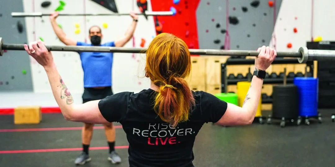 A woman with red hair, wearing a black shirt that reads RISE. RECOVER. LIVE., performs an overhead barbell press in a gym, with a man lifting a barbell in the background.