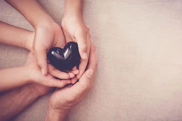Multiple pairs of hands cupped together holding a smooth, black heart.