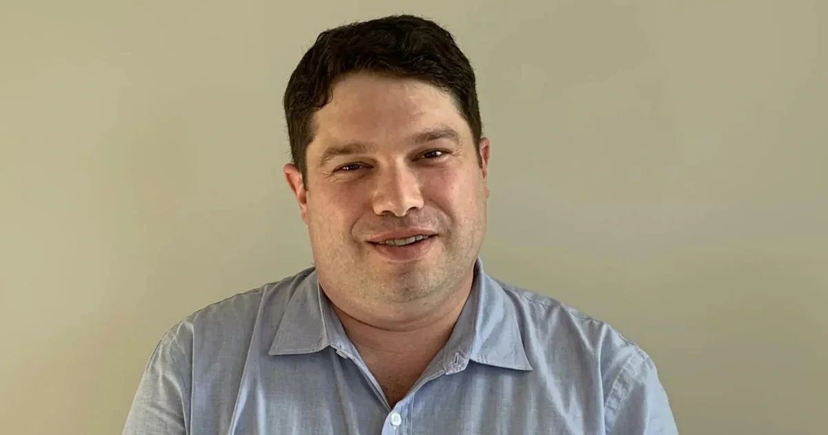 A man with dark hair wearing a light blue button-down shirt.