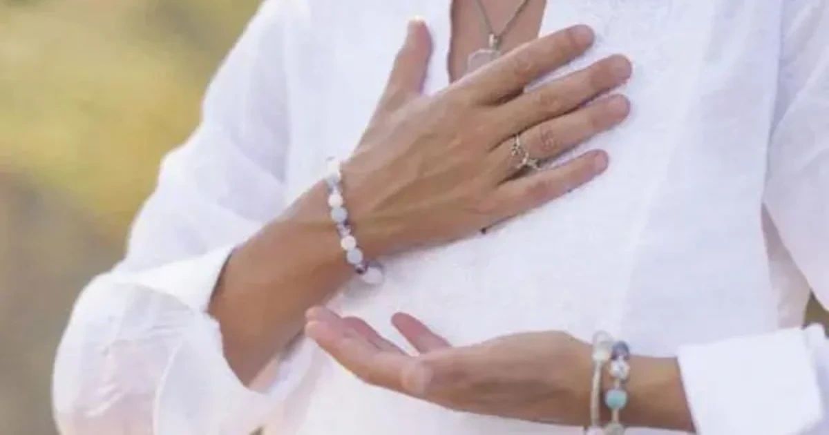 A person wearing a white shirt and beaded gemstone bracelets holds one hand to their chest with the other hand open, palm up.
