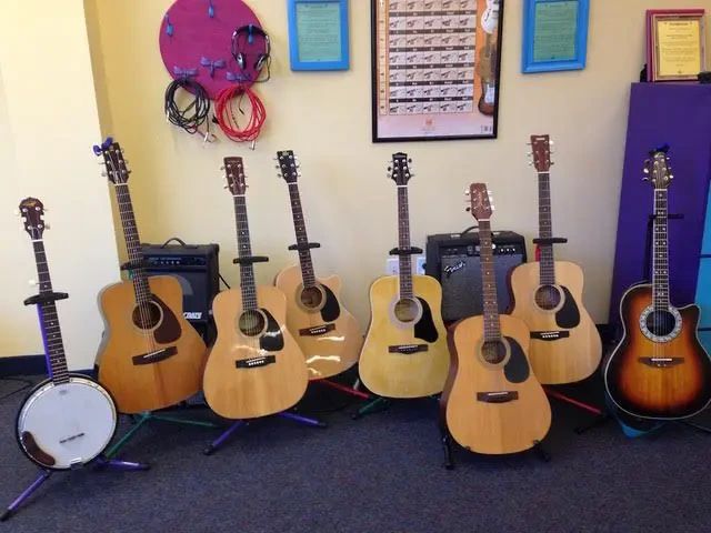 A collection of seven acoustic guitars and one banjo on stands, arranged in a row in a room with posters and amplifiers.