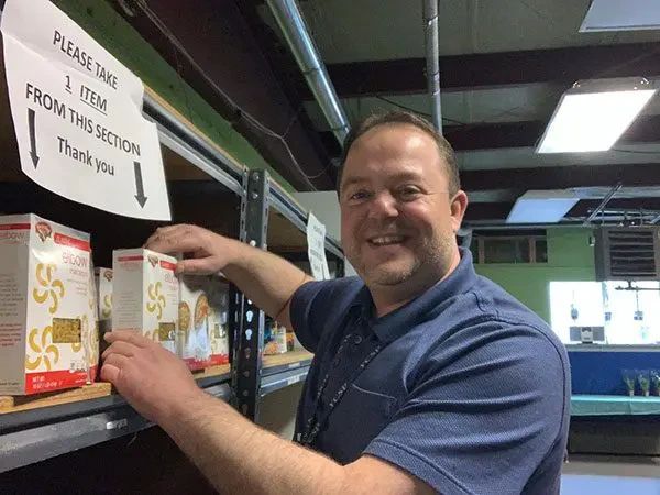 A smiling man in a blue polo shirt reaches for a box of pasta on a metal shelf next to a sign that reads, Please take 1 item from this section.
