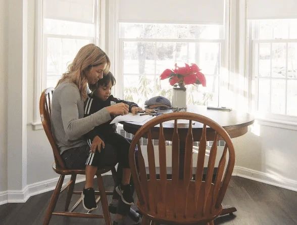 A woman sits at a table with a young child on her lap, helping them draw with crayons.