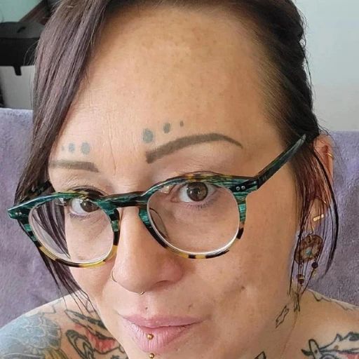 A person with glasses, facial piercings, and several tattoos is looking at the camera. Embodying a unique journey, Crystale Vega has drawn or tattooed dots above their eyebrows and is wearing earrings.