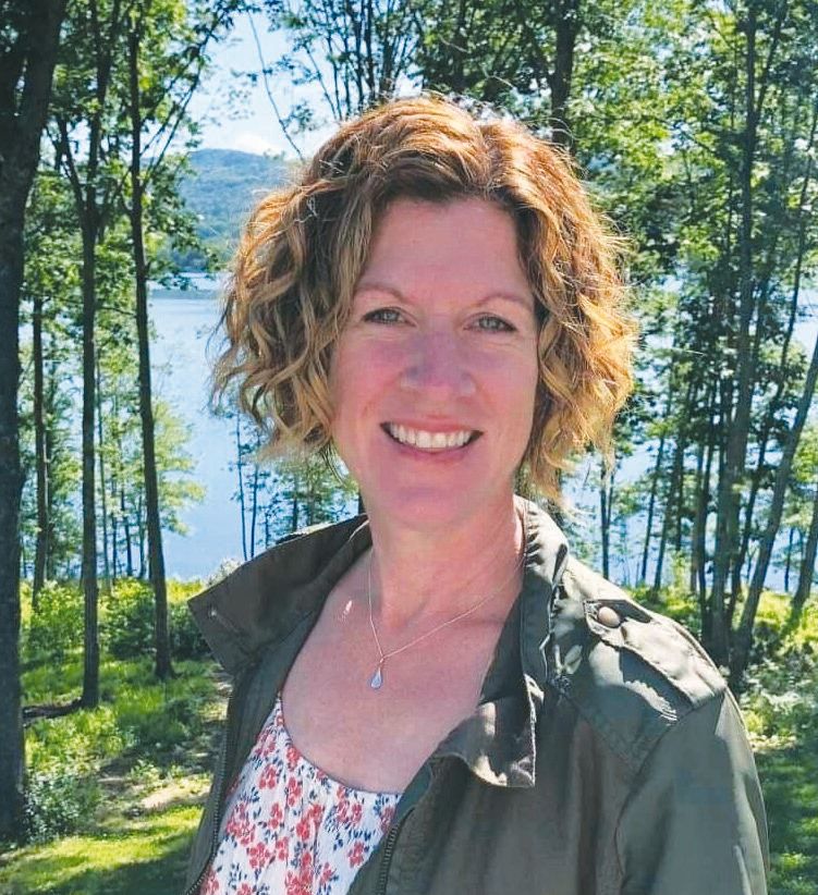 A woman with curly, short hair smiles outdoors in Maine, wearing a floral top and green jacket. Behind her are tall trees, green foliage, and a lake with hills in the background—a bright day captured by Maine Voices of Recovery.