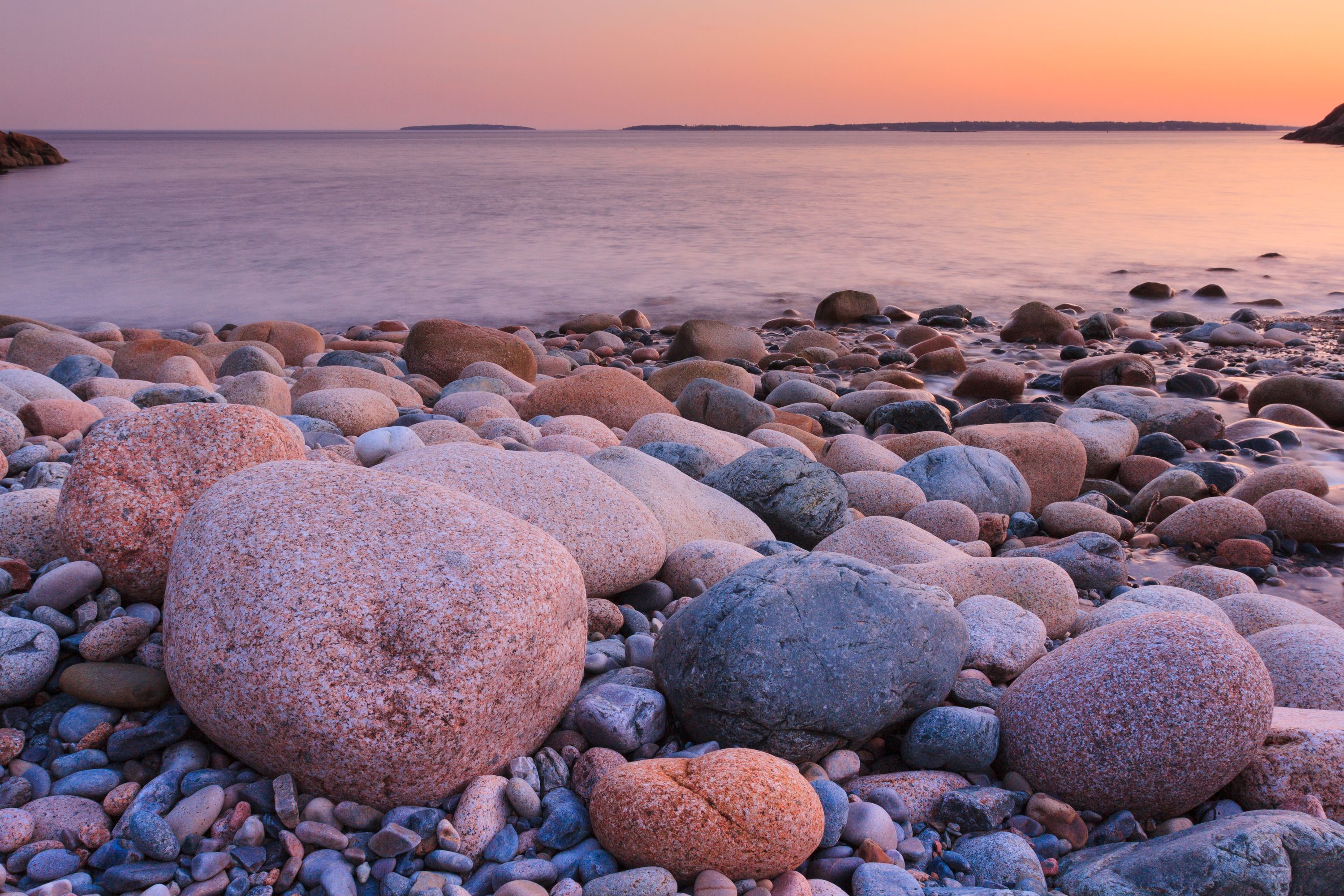 Smooth, rounded stones line a beach at sunset, with a calm ocean under a soft pink and orange sky.