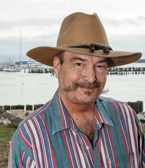 A man wearing a tan cowboy hat and a multicolored striped shirt, standing in front of a body of water with boats in the distance.