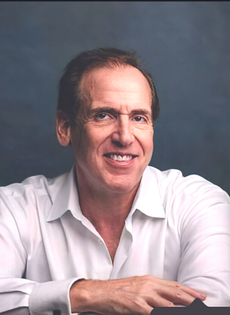 Brian Cuban, smiling, in a white open-collar shirt against a slate-blue background.