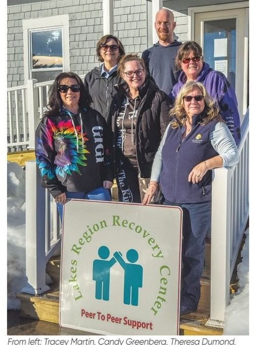 Six people standing on a porch holding a Lakes Region Recovery Center sign.
