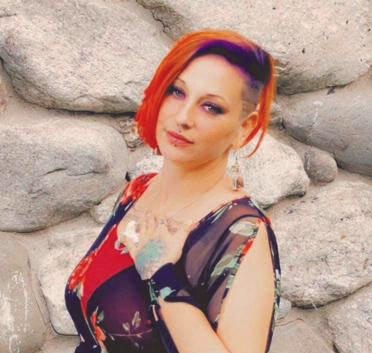 Christine Maider, with vibrant red and purple hair, wearing a floral dress and earrings, stands before a wall of large gray stones. She poses with one hand on her chest, looking at the camera. The image appears pixelated.