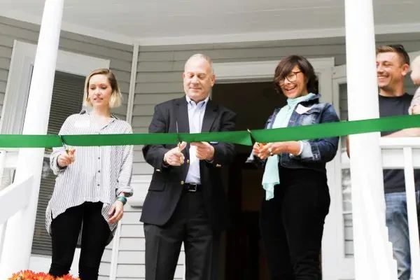 Three people cut a green ceremonial ribbon in front of a house entrance.