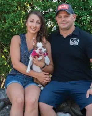 A woman and a man sitting outdoors, holding a small dog wearing a pink bandana.