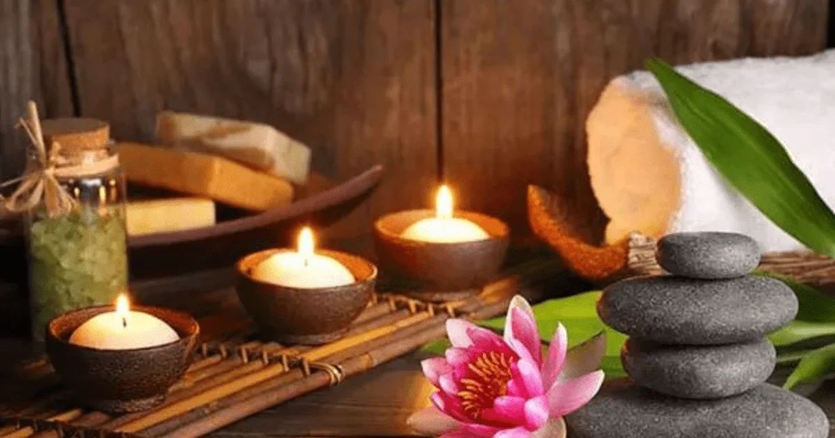 Three lit candles, a lotus flower, a stack of balance stones, and a rolled towel arranged on a wooden surface.