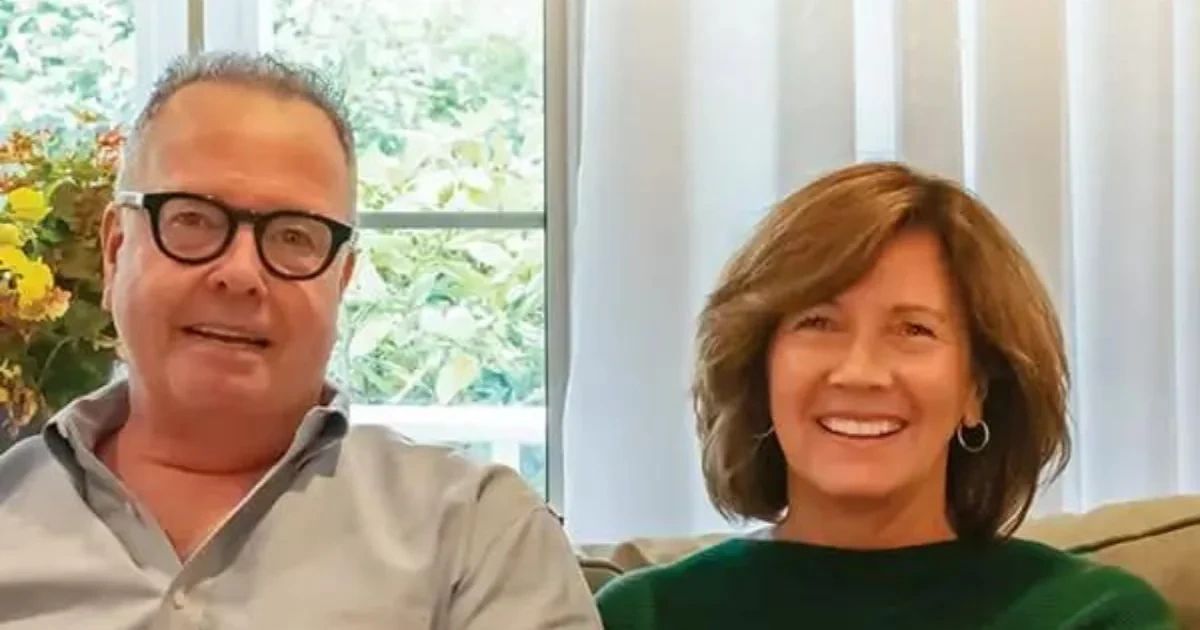 A middle-aged man wearing glasses and a light-colored button-down shirt sits next to a middle-aged woman with shoulder-length brown hair wearing a green sweater.