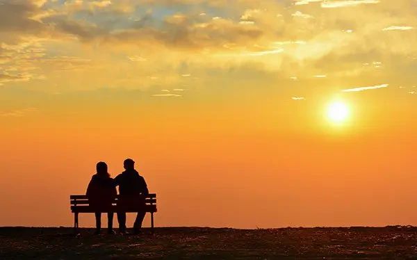 A couple sitting on a bench against a golden sunset.