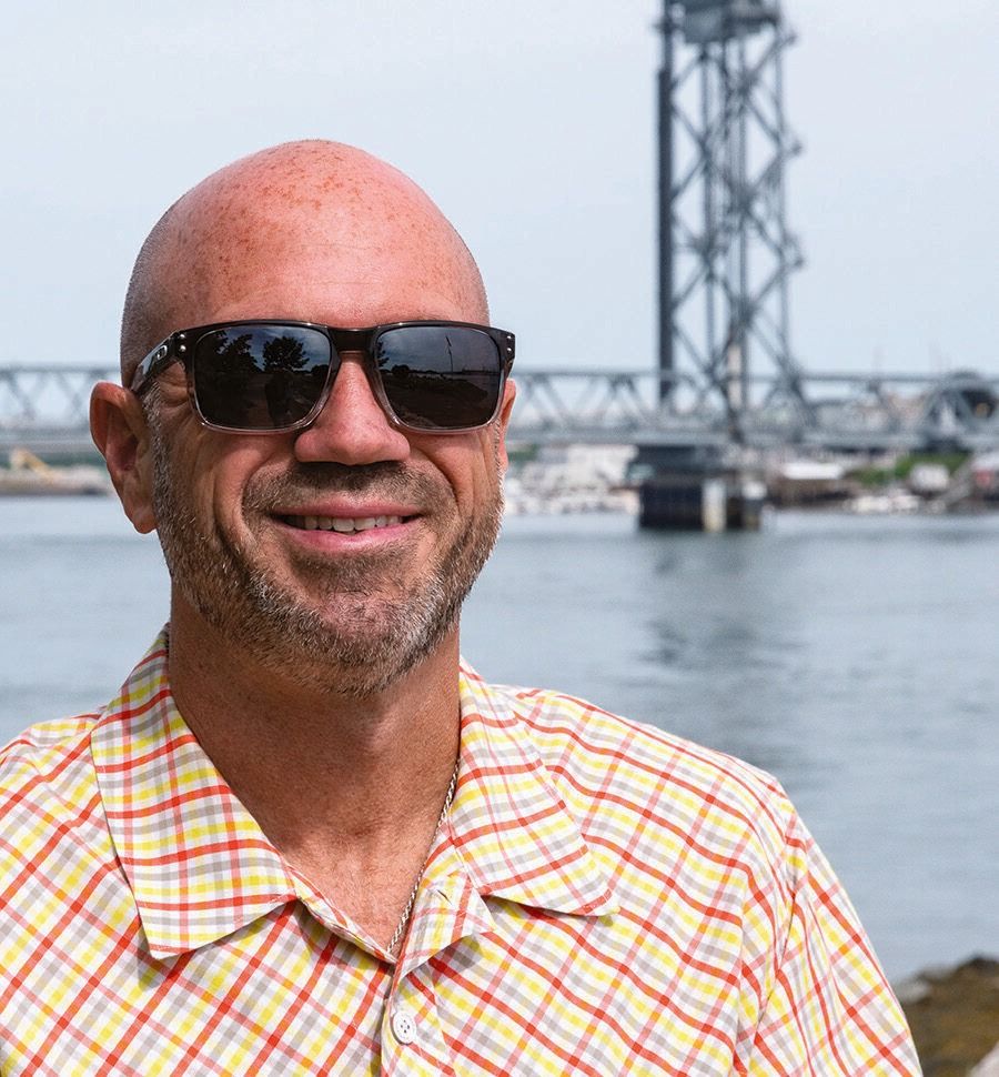 Freddy Petrone, a bald man wearing sunglasses and a yellow plaid shirt, smiles while standing near a body of water, with a bridge in the background on a bright day.