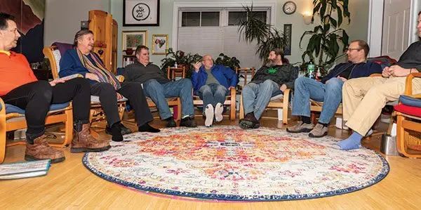 Six men sit in a circle on chairs in a living room, engaged in conversation around a large, colorful, patterned circular rug.