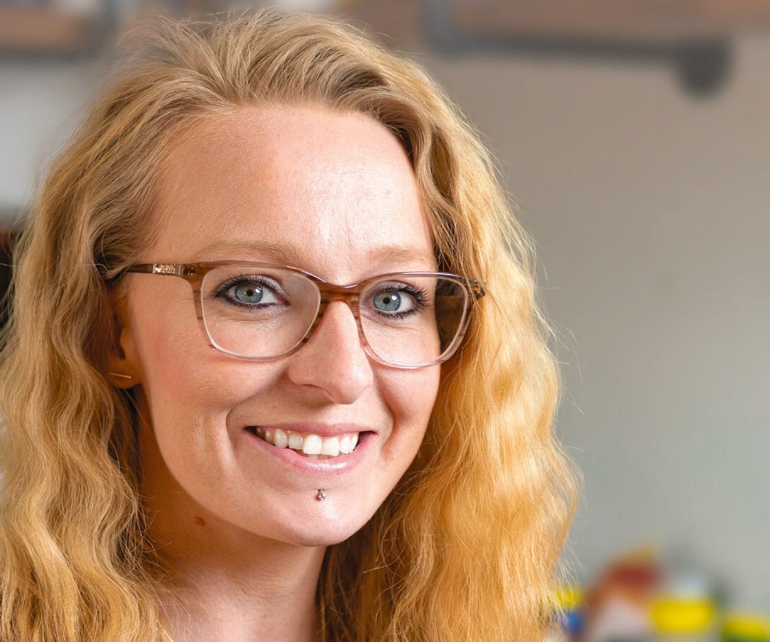 Sarah Langevin, a woman with long wavy blonde hair and glasses, smiles at the camera. She has a lip piercing and is in a brightly lit indoor setting.