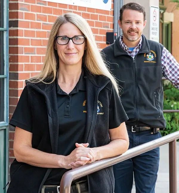 A woman and a man in matching Department of Correction uniforms stand together in front of a brick building.