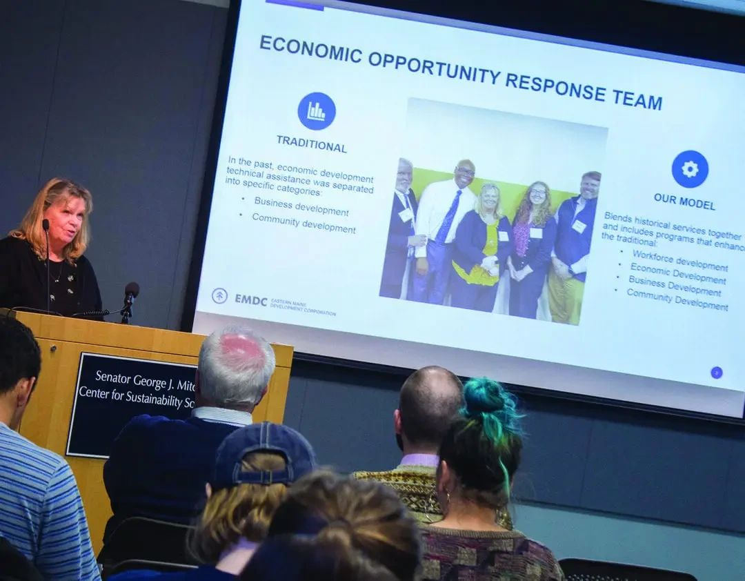A woman speaks at a podium in front of a projection screen displaying an Economic Opportunity Response Team presentation, with an audience seated before her.