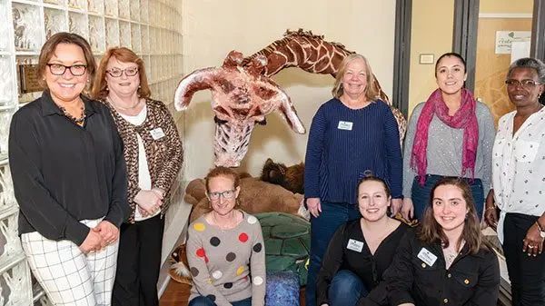 A group of seven women stand together in an office setting in front of a large giraffe sculpture.