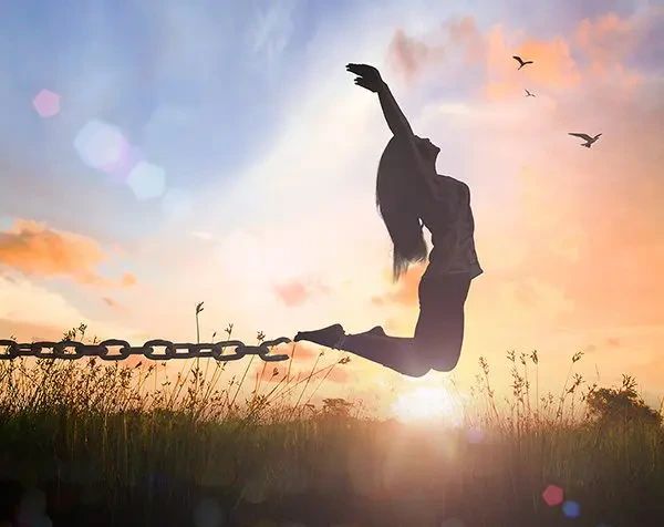 A silhouette of a person jumping with arms raised, breaking free from a heavy chain in a field at sunset.