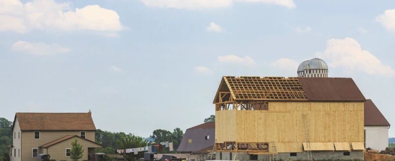 A partially constructed barn with exposed wooden framework stands beside a traditional farmhouse and silo under a partly cloudy sky—an image reflecting the strength of rural communities and the spirit of coalition building.