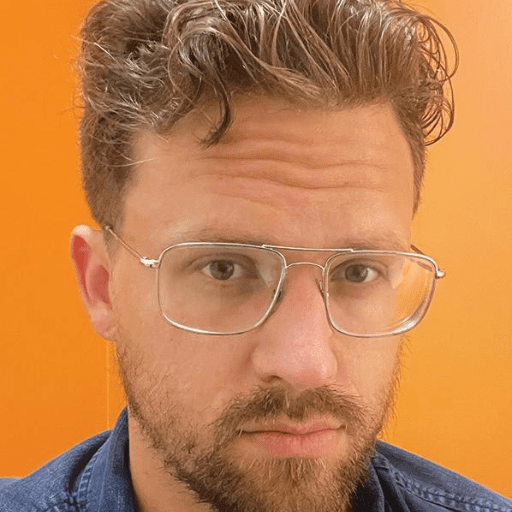 James Dillon III, a man with light brown hair and a short beard, wears clear-framed glasses and a blue shirt, standing in front of an orange background. He has a serious expression on his face.