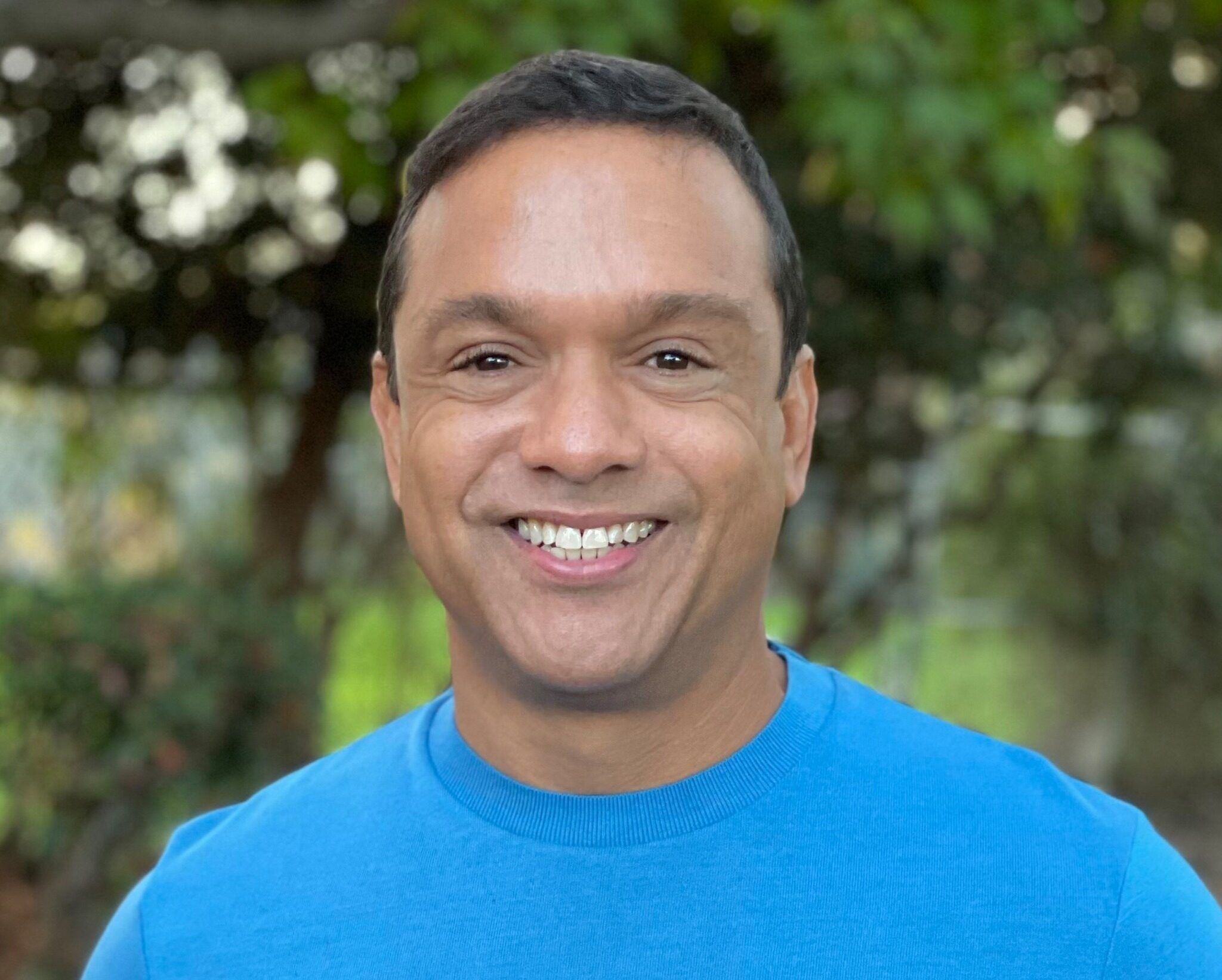 Kenny Williams, with short dark hair and a bright blue sweater, is smiling outdoors amid blurred green trees, embodying someone choosing to be of service.