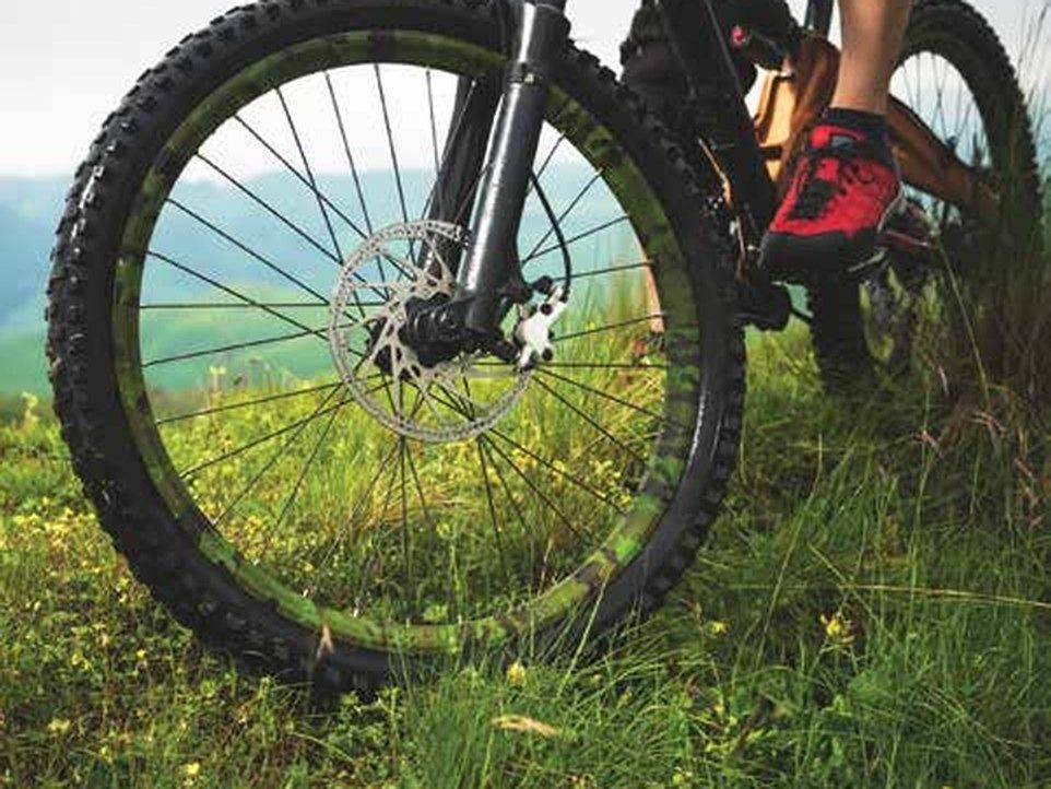 A mountain bike wheel with a camo-patterned rim resting in grassy terrain, with a cyclist’s foot in a red shoe visible on the pedal.