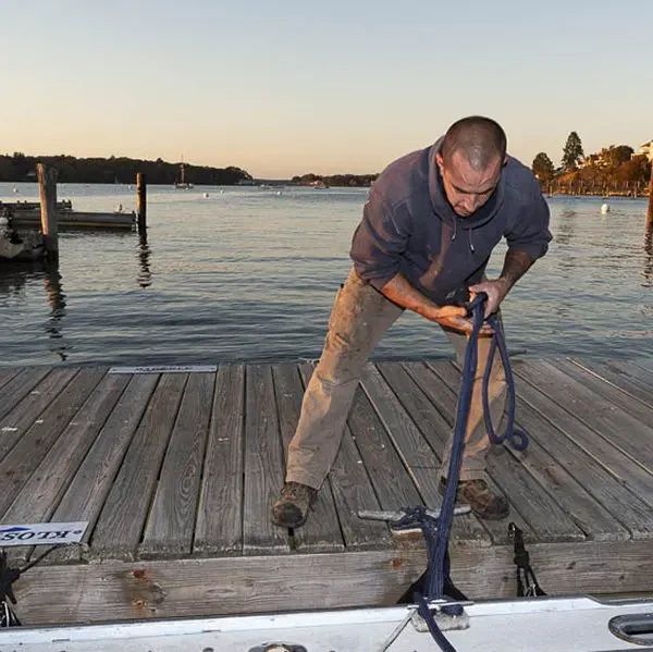 A man in a dark hoodie and work pants stands on a wooden dock, securing a blue rope to a metal cleat on a boat.