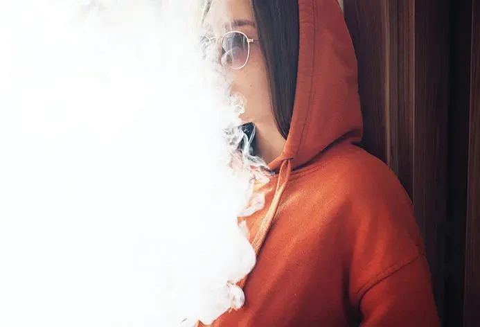 A person wearing a hooded sweatshirt and sunglasses exhales a large cloud of vapor.