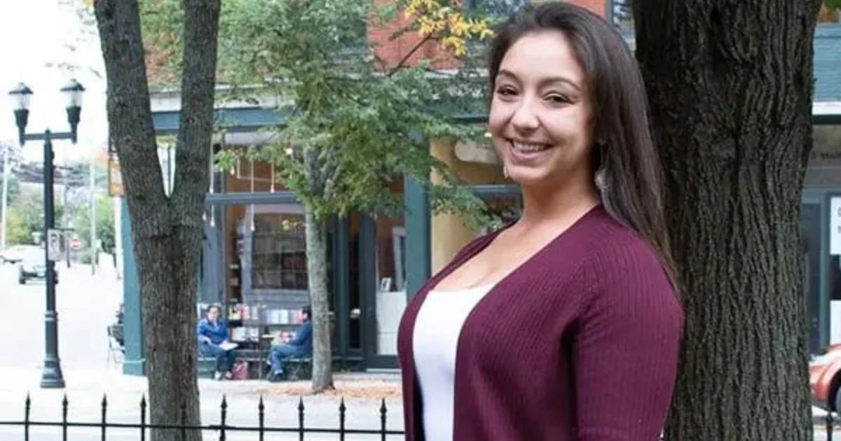 A smiling woman stands outdoors in front of a tree and a storefront with outdoor seating.