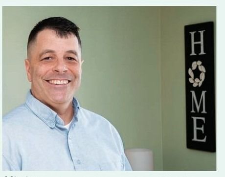 A smiling man in a light blue button-down shirt stands in front of a wall featuring a vertical sign that reads HOME with a wreath replacing the letter O.