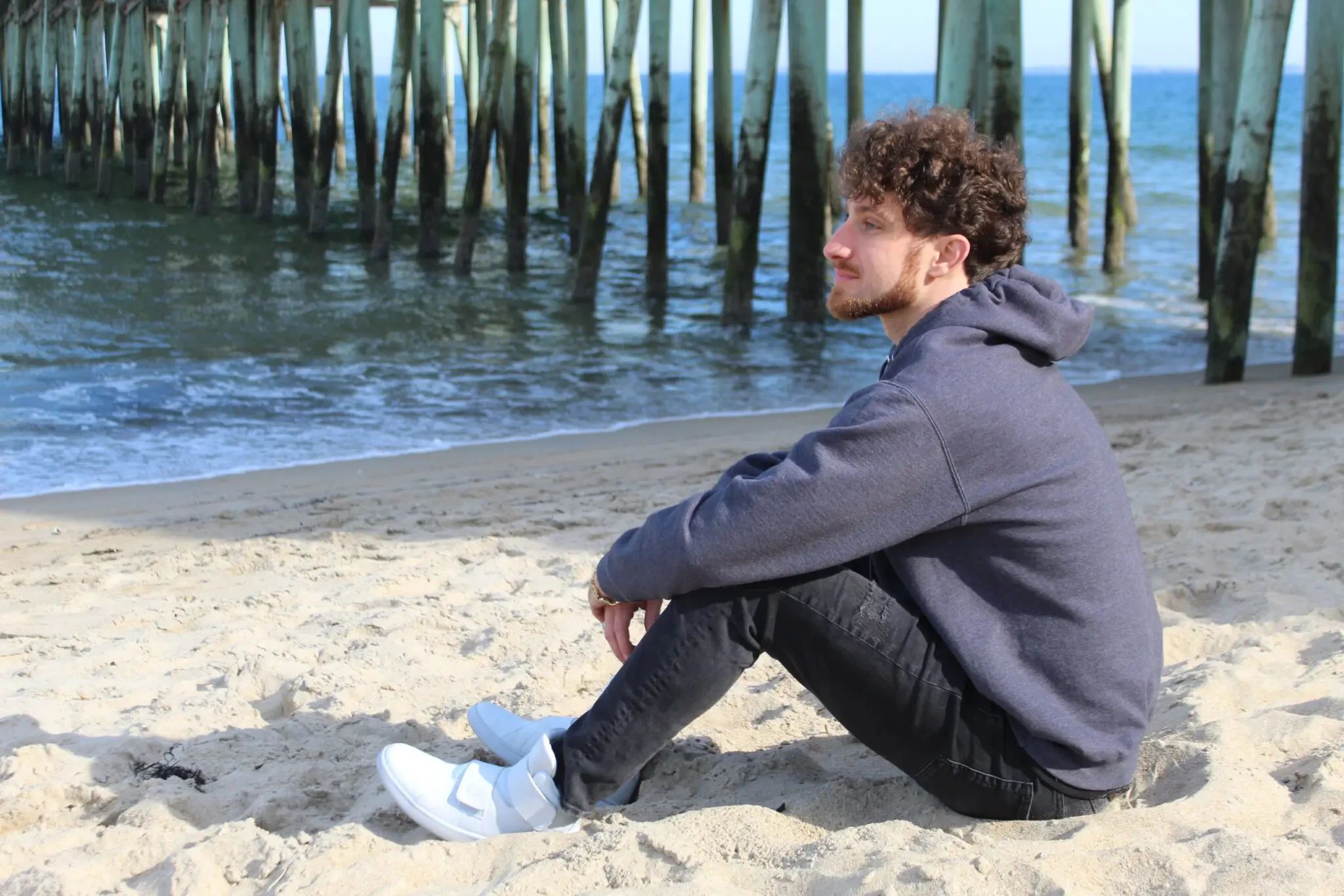 A man with curly hair and a beard sits on the beach, looking out toward the ocean near a wooden pier.